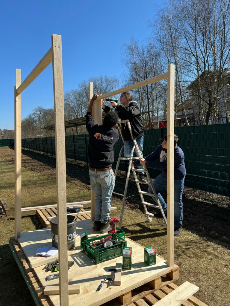 Mit einem Bohrer in der Hand steht eine Frau auf der Leiter und baut am gerüst der Kompsttoilette. Zwei Männer helfen ihr. 