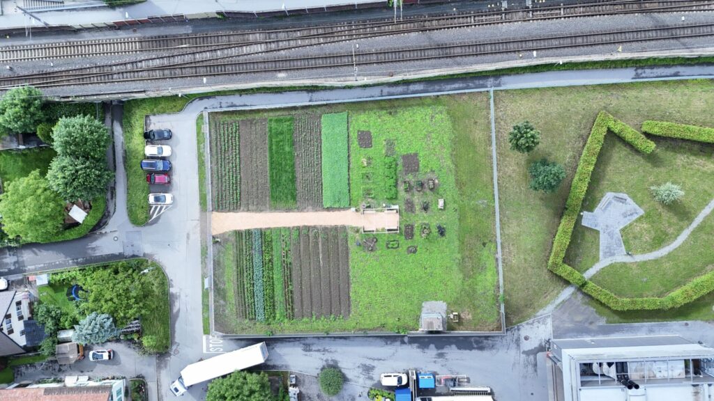 Aus der Luft ist der Weltacker Hohenems zu sehen. Oberhalb sind auf dem Bild Bahnschienen zu sehen, rund herum Straßen und Gebäude.