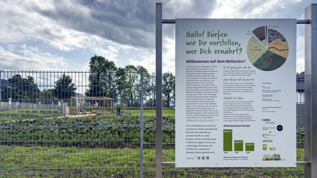 Vor einem Zaun ist das Eingangsschild mit dem Titel "Hallo! Dürfen wir Dir vorstellen, wer Dich ernährt?" angebracht. Im Hintergrund ist der Weltacker zu sehen.