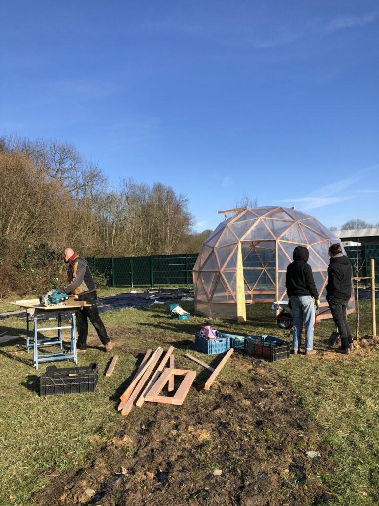 Ein Mann sägt Bretter für das Geodom zu - im Hintergrund ist das fast fertige Geodom zu sehen. 