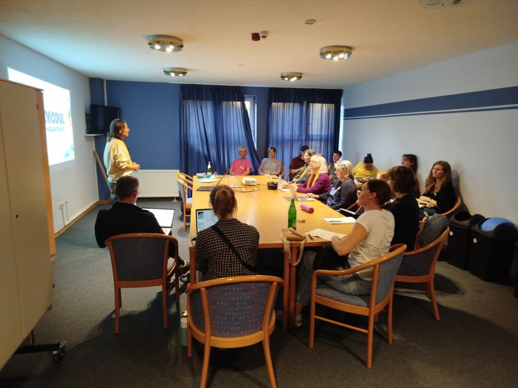 Rund um einen großen Tisch sitzen etwa 15 Menschen und lauschen der Vortragenden