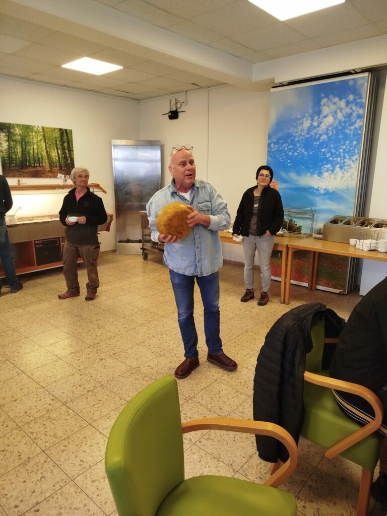 Ein Mann steht mit einem großen Käse in der Hand, andere Personen blicken ihn gespannt an.