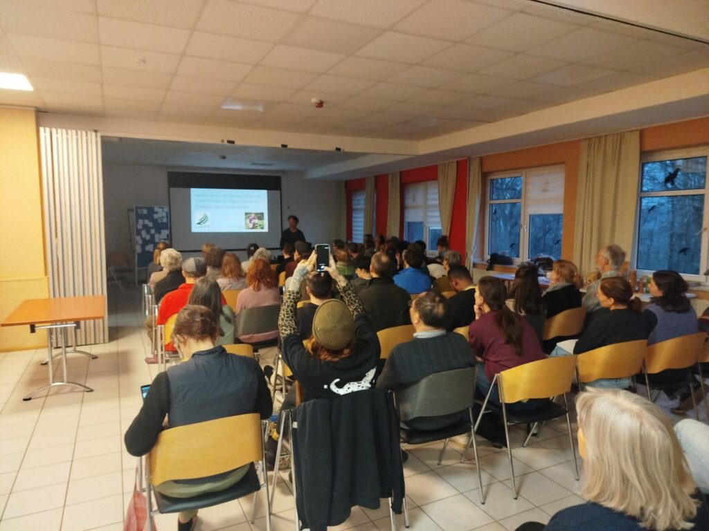 In einem Raum sitzen zahlreiche Menschen. Vorne erkennt man einen Mann, der eine Präsentation hält.