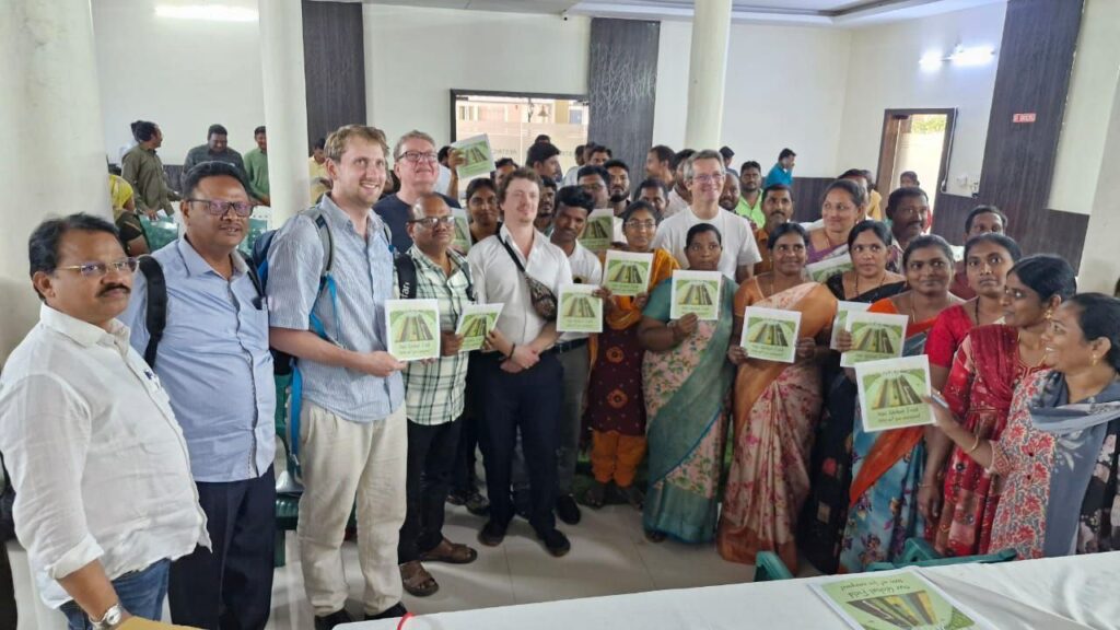 The Global Farmers delegation stands in a room surrounded by many Indian farmers. They are all holding Global Field brochures in the air.