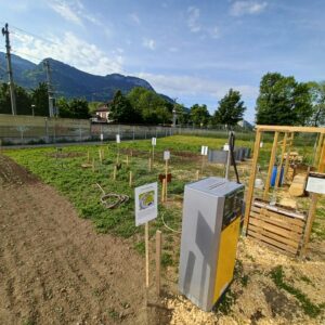 Auf dem Bild ist eine Zapfsäule zu sehen. Dahinter sind verschiedene Felder zu erkennen und im Hintergrund des Weltackers die Schallschutzmauer zu den Schienen und die Alpen.