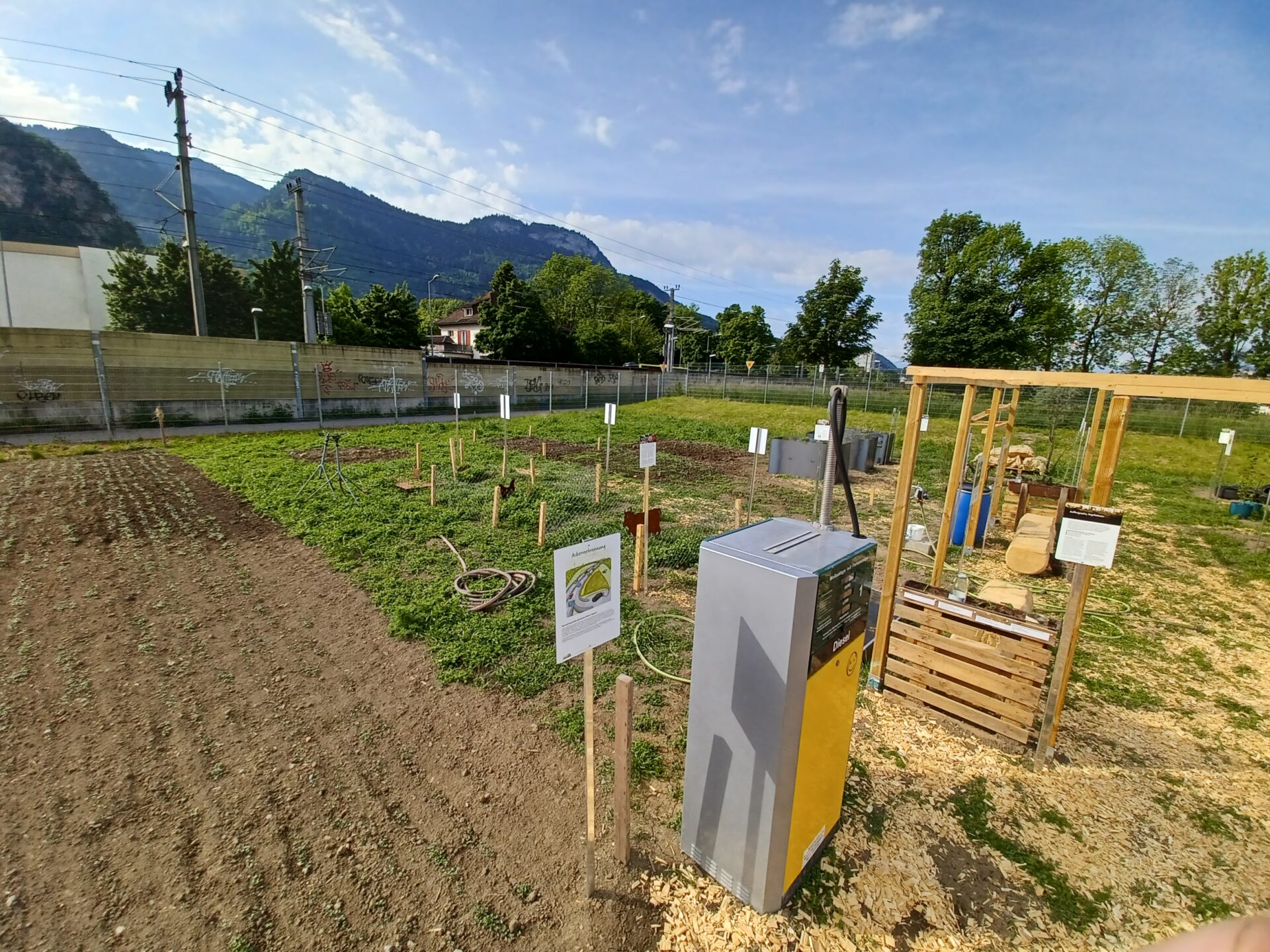 Auf dem Bild ist eine Zapfsäule zu sehen. Dahinter sind verschiedene Felder zu erkennen und im Hintergrund des Weltackers die Schallschutzmauer zu den Schienen und die Alpen.
