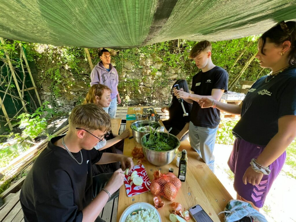 Fünf junge Erwachsene sitzen an einem Tisch und schneiden gemeinsam Gemüse. 