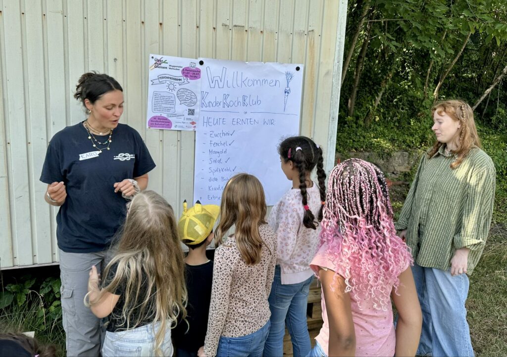Eine Gruppe Kinder lauscht der Bildungsreferentin aus Wuppertal, an der Wand hängt ein Plakat mit der Aufschrift "Willkommen im KinderKochKlub".